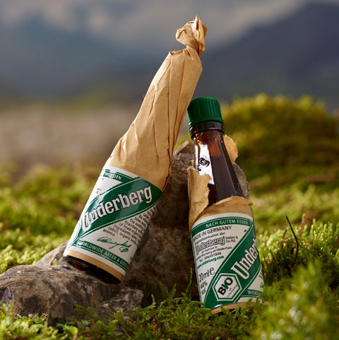 Two Underberg bottles with their labels peeled back, lying on grass with a mountainous background.